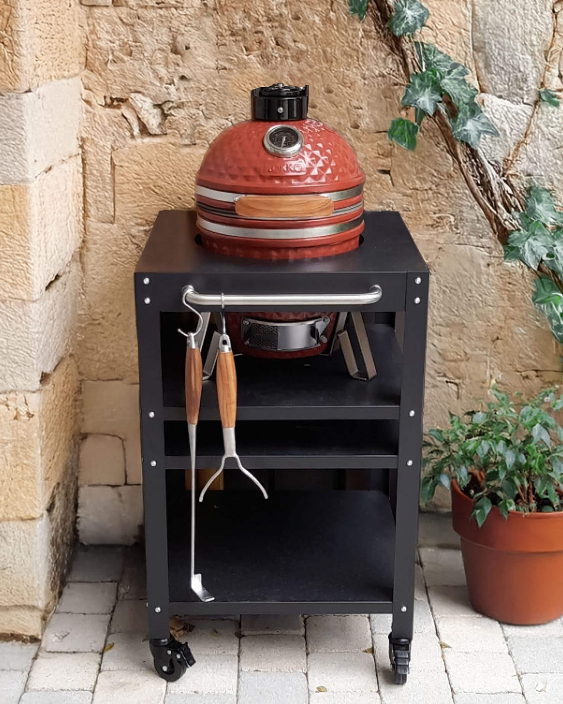 Kamado Kokko rouge en céramique installé sur un chariot noir à roulettes, avec tablette supérieure, espace de rangement ouvert et ustensiles suspendus à l’avant, présenté dans une cour en pierre.