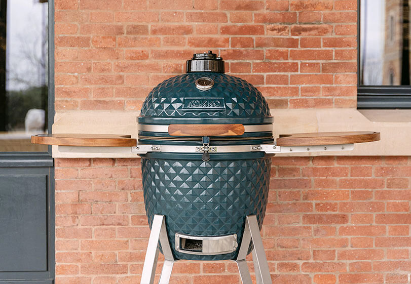 Kamado Kokko bleu océan installé sur support avec tablettes en bois, présenté de face en extérieur contre un mur en briques.