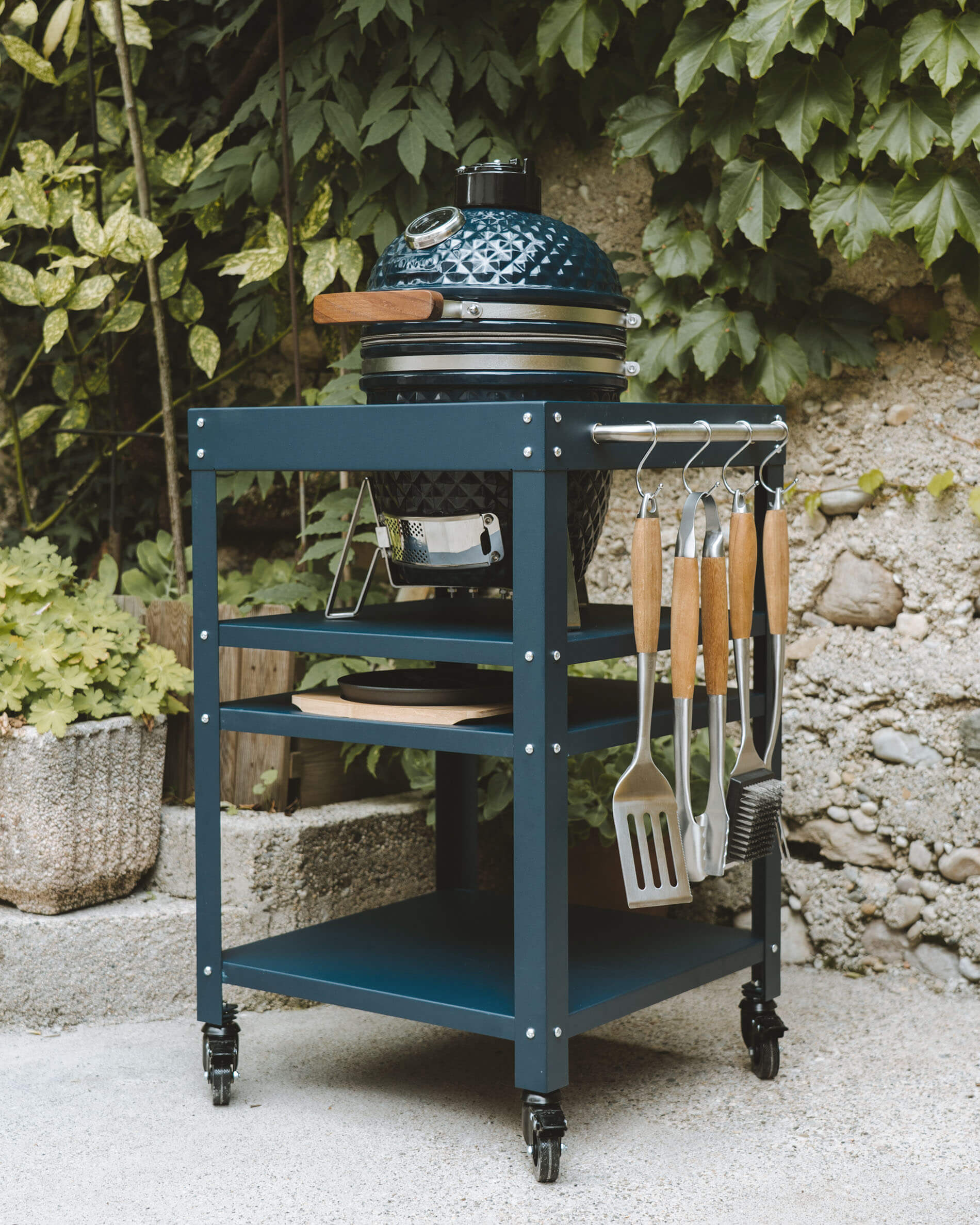Kamado Kokko coloris bleu océan installé sur une desserte métallique assortie, avec étagères de rangement, bac à cendres et ustensiles suspendus sur le côté.