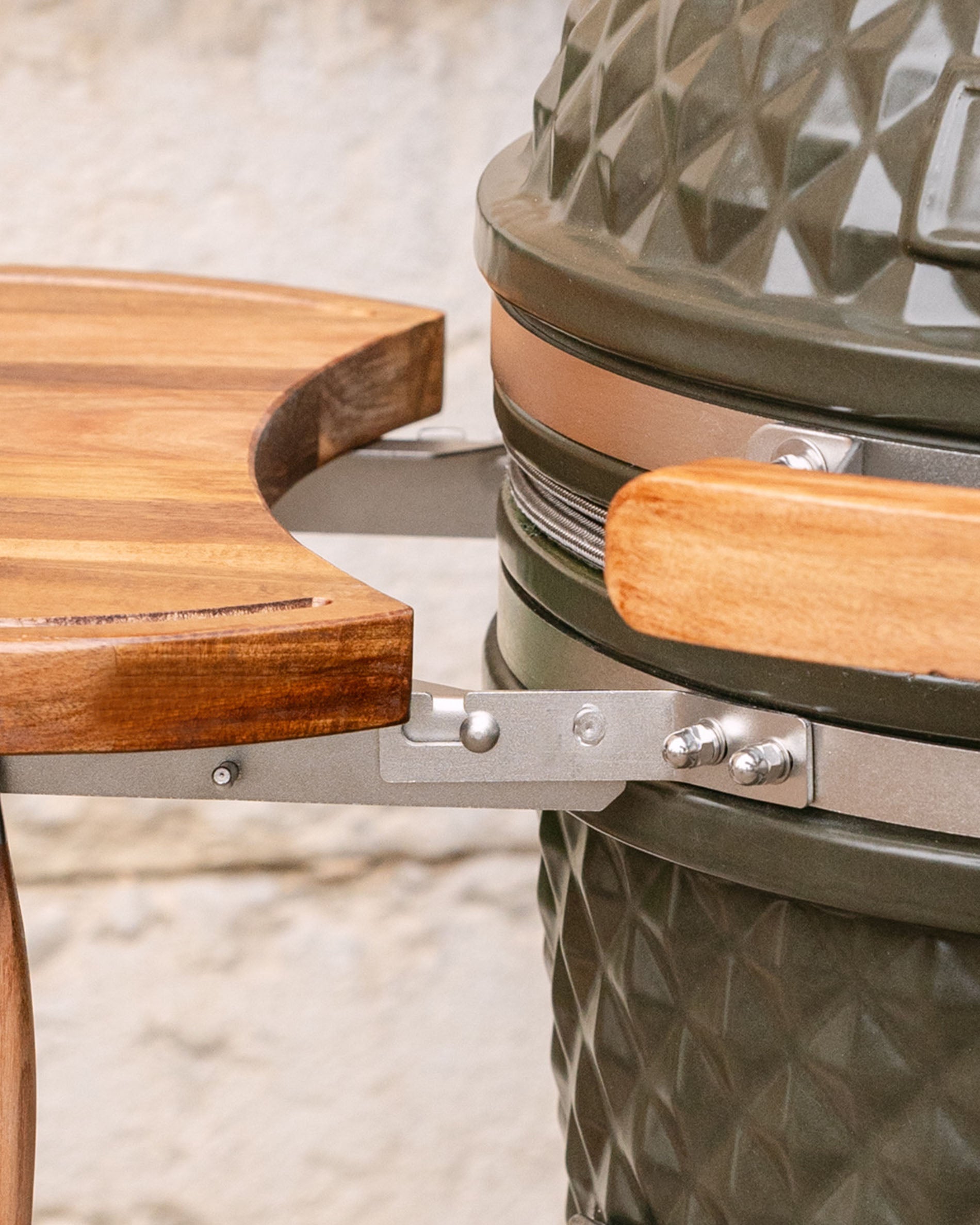 Détail des fixations métalliques reliant le plateau latéral en bois au kamado, avec poignée en bois et cerclage inox visibles.