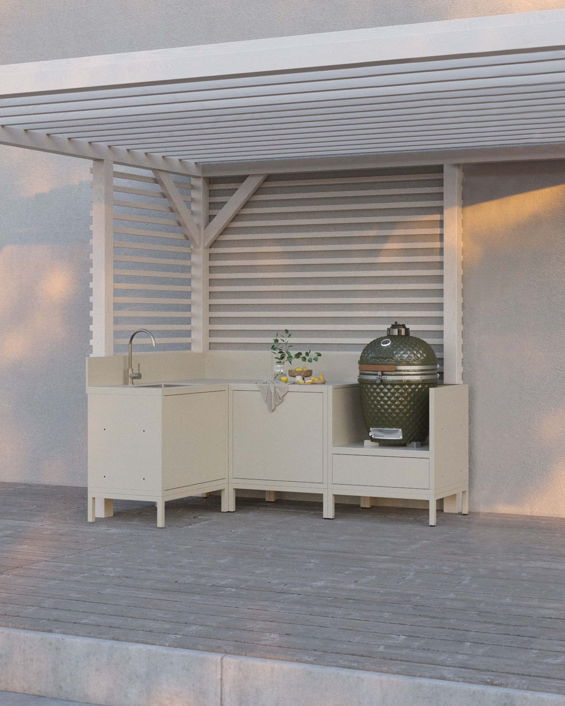 Cuisine pilotis coloris crème composée de quatre modules avec angle et évier, plan de travail continu et emplacement dédié au kamado, installée sous une pergola blanche sur terrasse en bois.